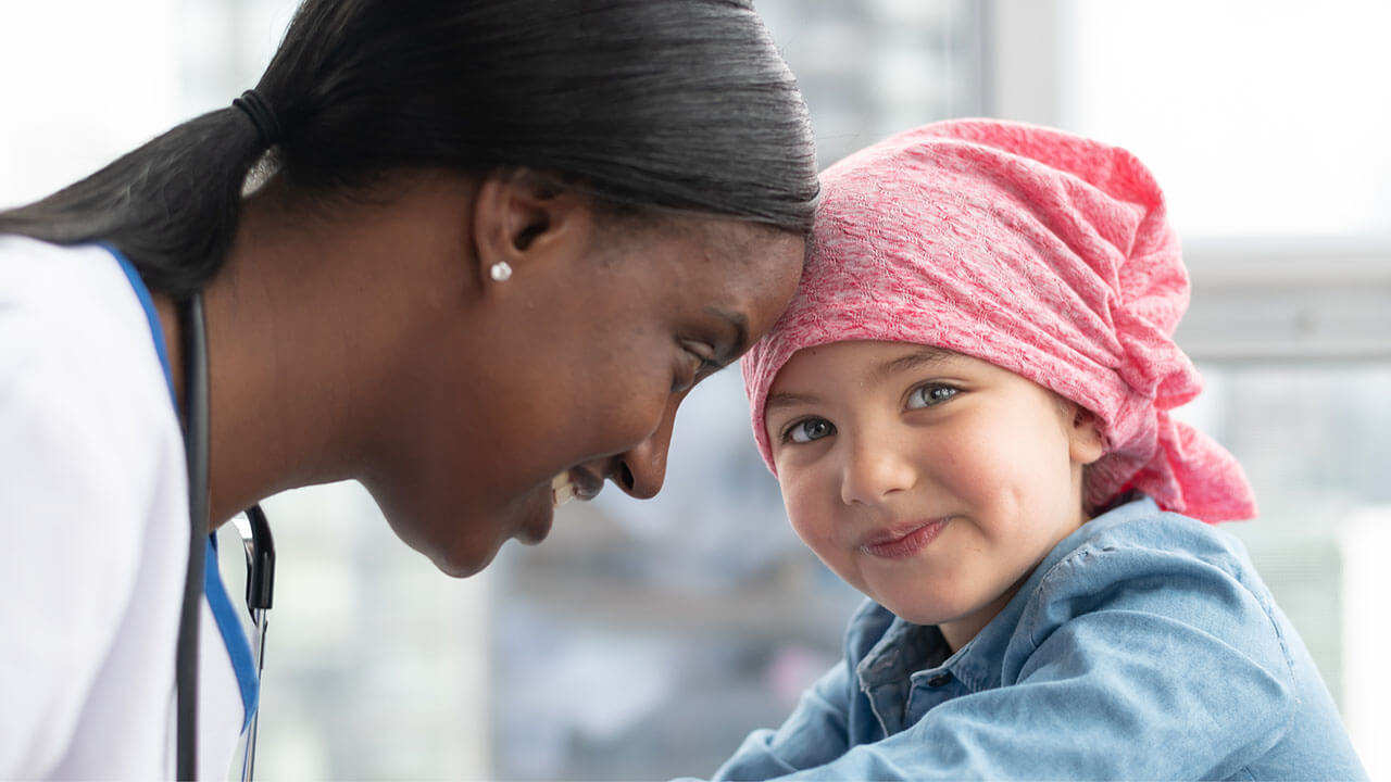 A female nurse leans her head against a child cancer patient's head