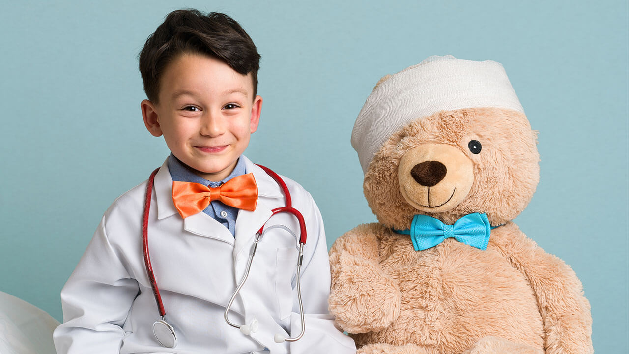 Young boy dressed as a doctor sitting next to a large teddy bear with a bandage on it's head